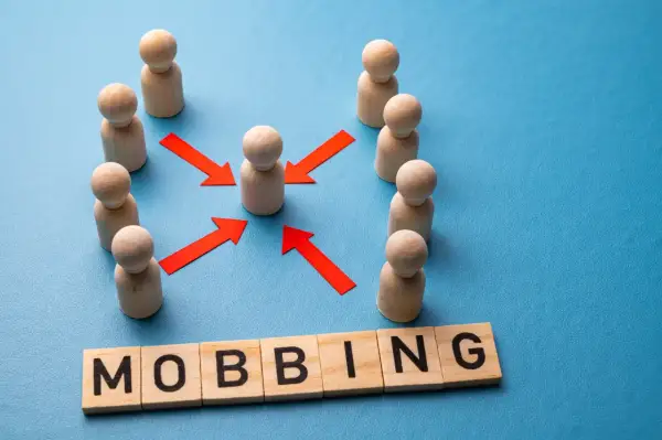 Drewniane pionki otaczają jednego, a czerwone strzałki wskazują na niego. Słowo "MOBBING" ułożone z klocków. Jak udowodnić mobbing w pracy?