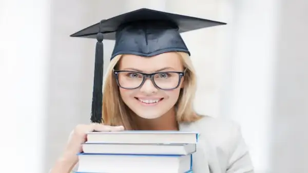 Uśmiechnięta studentka w birecie i okularach trzyma stos książek, zastanawiając się, ile trwają studia podyplomowe.