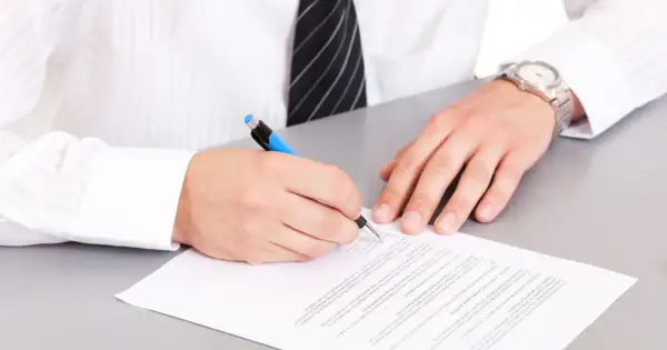 Mężczyzna w białej koszuli i krawacie podpisuje dokument. Na papierze widoczna jest klauzula CV po angielsku.