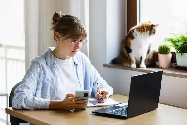 Kobieta w okularach pracuje zdalnie okazjonalnie, notując coś w zeszycie i patrząc na telefon. Kot obserwuje świat za oknem.