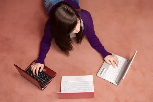 Kobieta leży na dywanie i pisze na trzech laptopach.