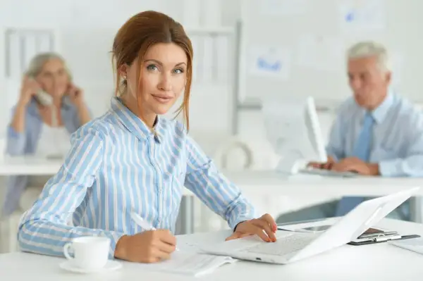 Kobieta na kursie księgowości pracuje przy laptopie, notując coś na papierze. W tle widać innych pracowników biurowych.