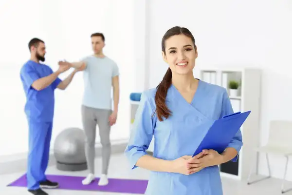 Uśmiechnięta asystent medyczny z tablicą w ręku, w tle fizjoterapeuta pracuje z pacjentem.