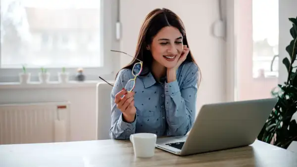 Uśmiechnięta kobieta z okularami w dłoni pracuje przy laptopie. To może być jej dzień w dziale **human resources**.