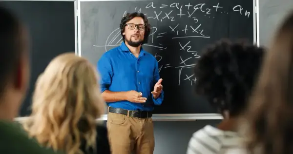 Nauczyciel w niebieskiej koszuli tłumaczy matematykę przy tablicy. Czy nauczyciel jest funkcjonariuszem publicznym? To pytanie nurtuje wielu.