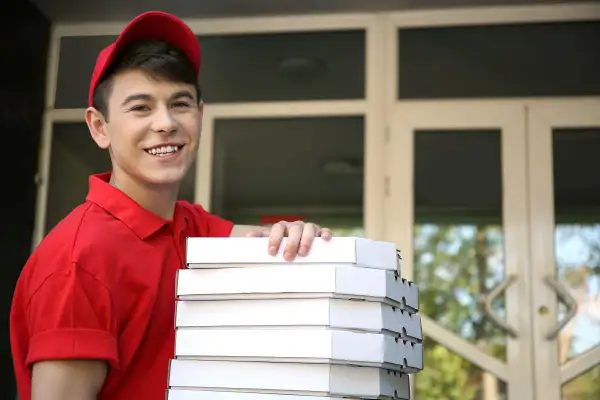Uśmiechnięty młodociany dostawca pizzy w czerwonej koszulce i czapce trzyma stos pudełek z pizzą przed drzwiami.