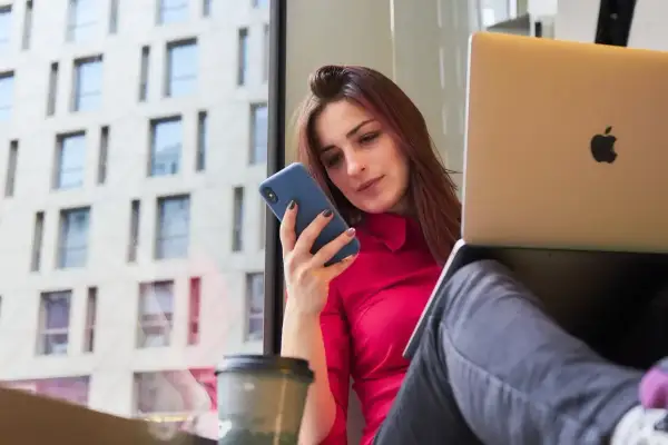 Kobieta w czerwonej bluzce korzysta z telefonu i laptopa, pracując w domu.