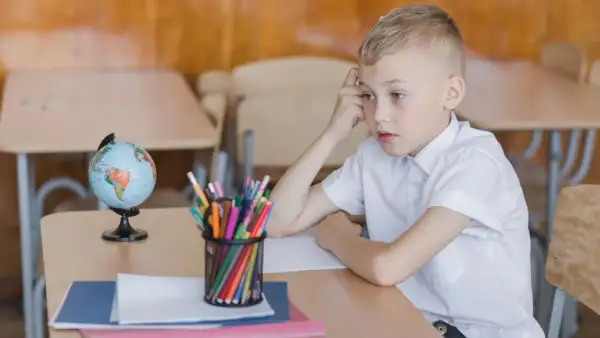Chłopiec zamyślony przy biurku, na którym leży globus i przybory. Obowiązek szkolny bywa trudny.
