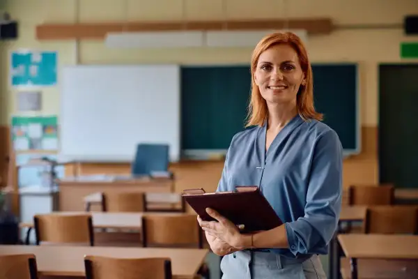 Uśmiechnięta nauczycielka z teczką w ręku, w tle klasa szkolna. Legitymacja nauczyciela to symbol jej pracy i zaangażowania.