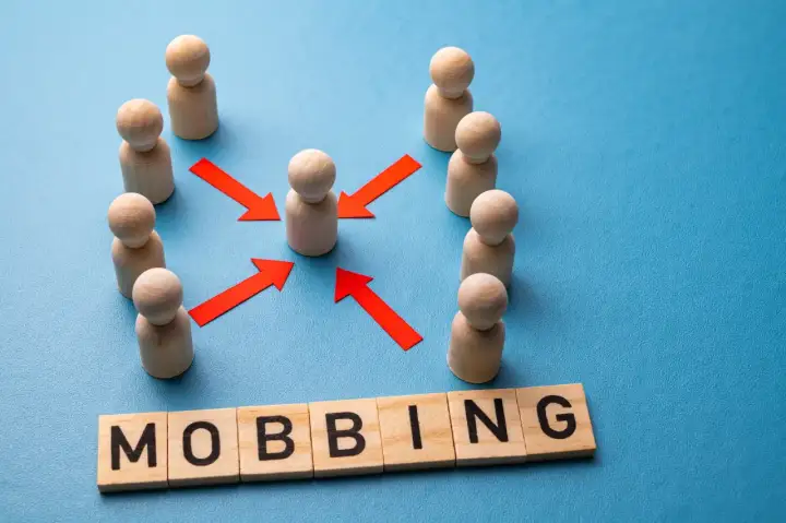 Drewniane pionki otaczają jednego, a czerwone strzałki wskazują na niego. Słowo "MOBBING" ułożone z klocków. Jak udowodnić mobbing w pracy?